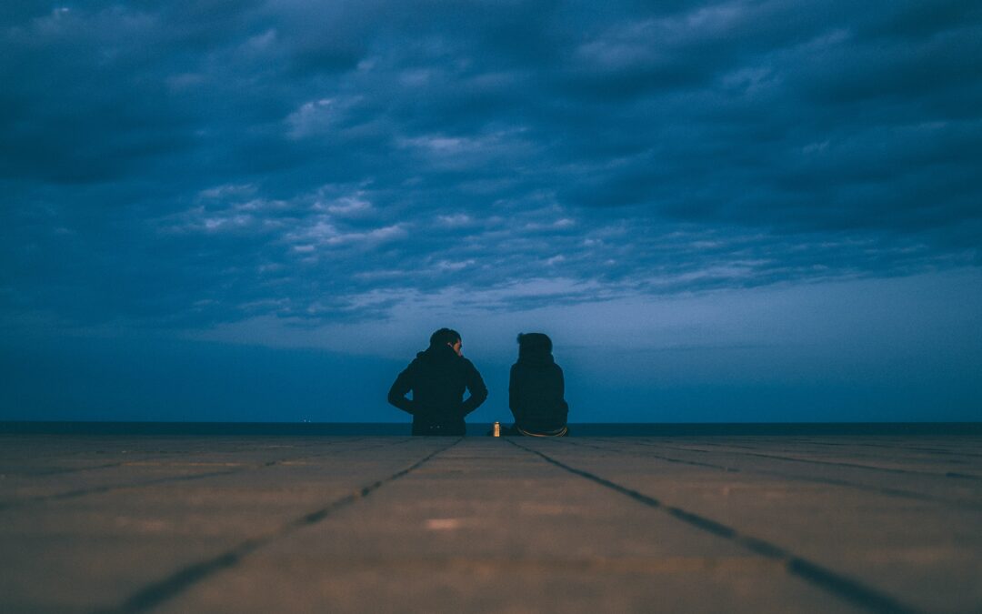 Two people sitting as the sky gets dark having a conversation. Their back is turned to the camera.